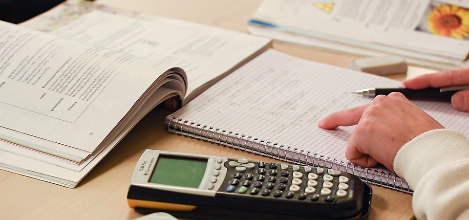 En närbild på där någon räknar matematik med en miniräknare bredvid sig. Ett finger pekar på ett block. 